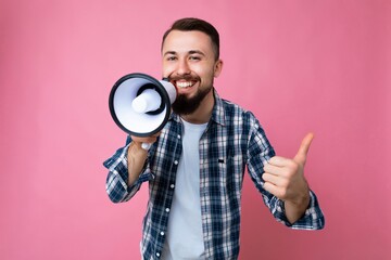 Photo of young handsome positive happy smiling brunet man with beard with sincere emotions wearing t-shirt and stylish check shirt isolated over pink background with copy space and speaking into