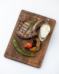 Grilled Meat steak ribeye. Tomahawk or t-bone steak with grilled vegetables and sauce on wooden cutting board. Object isolated on white background. Top view. Vertical format.