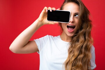 Photo of beautiful smiling young blonde woman good looking wearing white t-shirt standing isolated on red background with copy space holding phone showing smartphone in hand with empty screen display