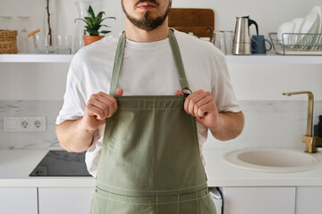 A man in a kitchen apron stands in a modern kitchen. Cooking at home in uniform, protection apparel. Green fabric apron, casual clothing. A man in an apron prepares to prepare a meal - obrazy, fototapety, plakaty