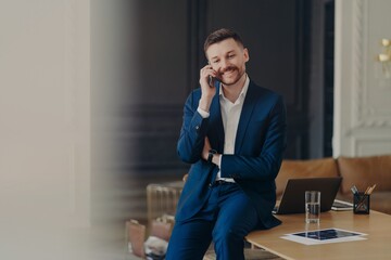 Happy handsome businessman talking on phone while sitting on office desk