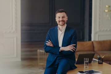 Photo of satisfied bearded prosperous businessman stands self assured in own cabinet or contemporary office keeps arms folded dressed formally smiles pleasantly satisfied with results of his work