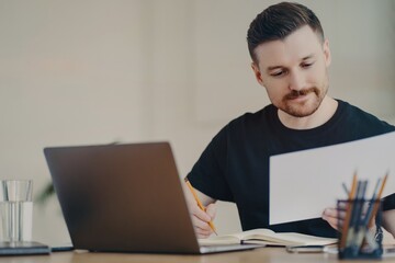 Skilled professional male expert busy doing paper report edits online files via laptop works at desktop focused at documents dressed in casual black t shirt. Bearded freelancer at home office