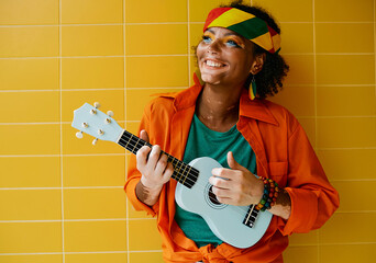 Fun rastafarian woman playing blue ukulele near yellow wall. Female street musician