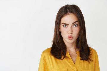 Close-up of confused and intrigued young woman raising an eyebrow, pucker lips and staring at camera puzzled, standing over white background