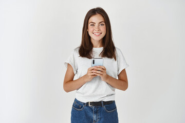 Image of smiling brunette female holding smartphone and looking at camera, standing in casual...