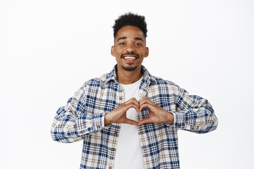 Stylish african american man smiling, showing heart on chest gesture, I love you, confess that he likes loves someone, romantic guy speaks true feelings, white background