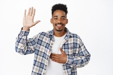 Greeting. Smiling friendly african american guy tell his name, raise hand and put arm on chest, introduce himself, say hello, making promise, standing over white background