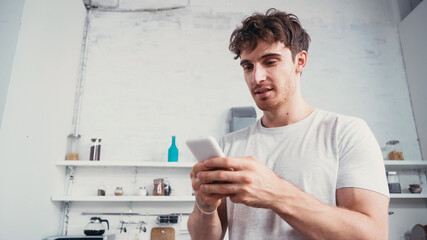 low angle view of young man in white t-shirt messaging on smartphone in kitchen