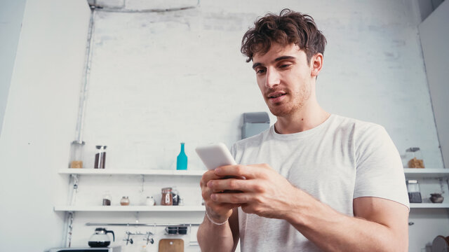 low angle view of young man in white t-shirt messaging on smartphone in kitchen - Powered by Adobe