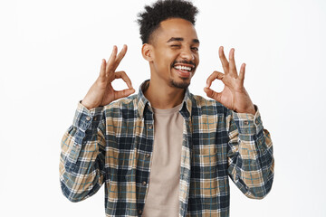 Fototapeta premium Perfect choice, okay. Smiling satisfied african american man, winking, nod in approval and show OK zero problems gesture, say yes, pleased something good, white background