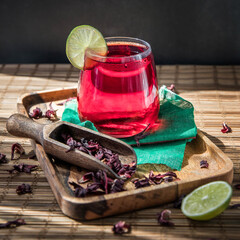 A glass of cold hibiscus tea on a wooden tray  and hibiscus tea brew .  A glass of red hibiscus tea on a green napkin .  An egyptian traditional drink .  Alternative tea without caffeine  © Eva