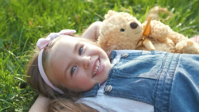 Pretty little child girl laying down with her teddy bear toy on blanket on green grass in summer smiling happily.