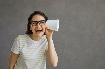 Young funny and curious woman on a gray background overhears a conversation with a plastic cup. Cheerful caucasian woman with a funny expression tries to hear what you are saying. Concept of curiosity