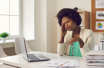 Tired exhausted young dark skin woman experiencing severe headache sitting at work in front of...