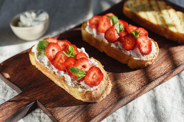 Two toasts or bruschetta with strawberry and cream cheese on wooden board. Summer breakfast. Hard lighting