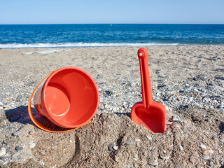 Fototapeta na wymiar Red plastic shovel and bucket for children in the sand on the beach in front of the sea, iconic image of summer and vacation period 