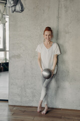 Young athletic ginger lady in activewear leaning against wall with fitball in hands