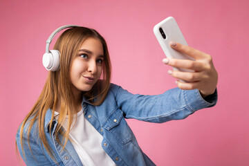 Photo of sexy beautiful happy adult blonde woman wearing blue jean shirt and white t-shirt isolated over pink background wearing headsets and listening to music and using mobile phone taking selfie - Powered by Adobe