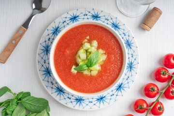 Top view of spanish gazpacho cold refreshing tomato soup made of raw blended vegetables eaten during hot summers served in bowl with spoon, basil leaves, cucumber cubes and glass of wine. Horizontal
