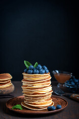 Pile of homemade sweet pancakes decorated with ripe fresh blueberries and green mint leaves topping served on brown round plate on dark wooden background for breakfast with fork and syrup. Vertical