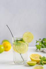 Sweet organic homemade lemonade summer cold drink made of sour ripe lemon and lime slices, sparkling water and mint leaves served in drinking glass with straw on white wooden table against gray wall