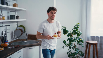 happy man in white t-shirt holding tasty pancakes with strawberry in kitchen