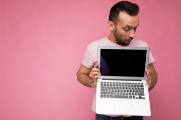 Handsome surprised and amazed man holding laptop computer looking at computer monitor in t-shirt on isolated pink background