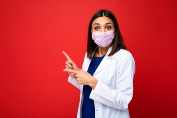 Amazed woman in virus protective mask on face against coronavirus and white medical coat isolated on the background and pointing fingers at copy space for information