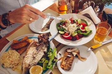 Man eats fish with vegetables and greek salad in outdoors restaurant. Fresh seafood dinner.