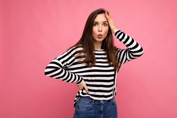 Photo of young astonished surprised beautiful brunette woman with sincere emotions wearing casual striped pullover isolated on pink background with copy space