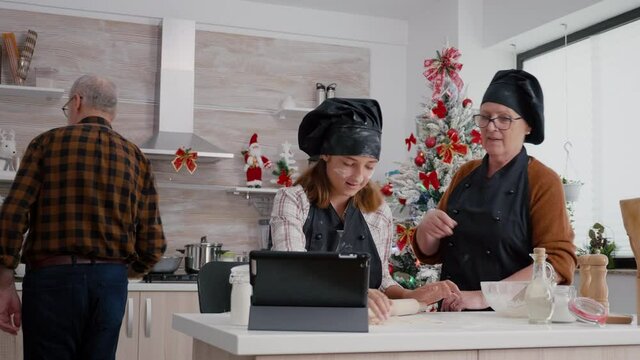 Granddaughter watching cook lesson using tablet computer preparing homemade gingerbread dough using rolling pin. Happy family enjoying cooking xmas dessert celebrating christmas holiday