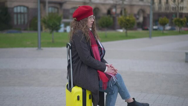 Young slim stylish Caucasian woman in red beret and eyeglasses sitting on yellow travel bag waiting. Portrait of dissatisfied elegant beautiful tourist in urban city outdoors. Tourism and lifestyle