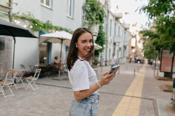 Cute lady in white top and jeans smiles widely and looks into camera. Happy brunette woman in good mood walks outside and listening music