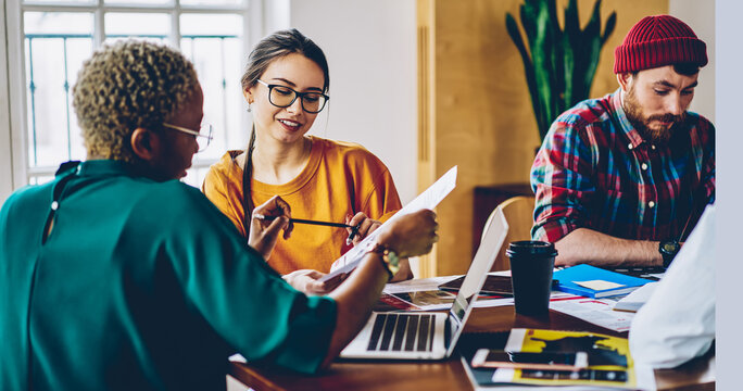 Carefree Caucasian employee in optical spectacles for provide eyes correction talking with dark skinned colleague discussing paperwork during collaborative briefing at desktop, business concept