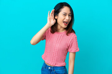 Young Vietnamese woman isolated on blue background listening to something by putting hand on the ear