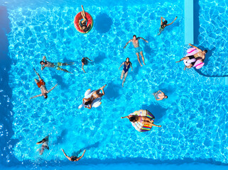 Aerial of friends having party in swimming pool with inflatable flamingo, swan, mattress. Happy young people relax at luxury resort on sunny day. View from above. Girls in bikini sunbathing in a sun.