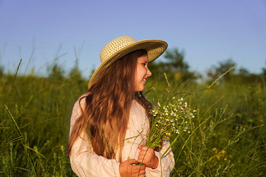 Portrait of cute smiling caucasian girl in a straw hat holding a bouquet of wildflowers walking in the meadow against of clear blue sky.  Summer sunset.  Happy Childhood concept.