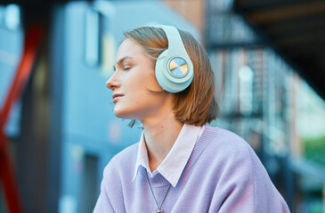 Melancholic lady student in earphones listens to music and looks around sitting on street against...