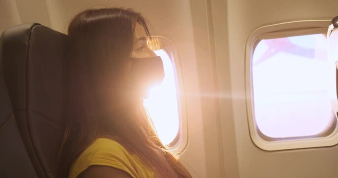 Close-up, young woman sits on airplane wearing medical mask near window. Head, shoulders of passenger. Sunset sun rays backlit traveler. New travel standards, normal. Traveling during COVID19 pandemic
