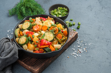 Fried vegetables in a frying pan on a gray background with fresh herbs.