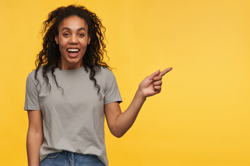 Indoor shot of young african american wears grey t-shirt, point with a finger aside into copy space, smiles broadly with positive facial expression. isolate over yellow background