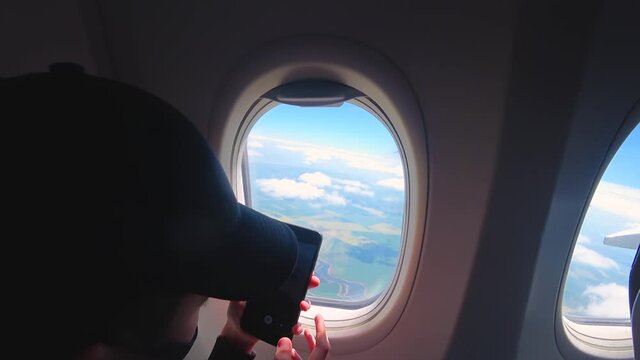 Silhouette of boy with mobile phone against background of airplane window. Child filming clouds on camera of smartphone by illuminator during flight in aircraft. Concept of trip to warm countries and - Powered by Adobe