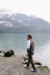 Fototapeta na wymiar A brutal thoughtful young man groom with a dreadlocked hairstyle in a fashionable elegant wedding suit stands on the shore of a lake against the background of misty mountains in nature