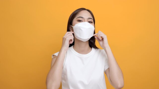 Young beautiful asian woman takes off mask after getting a vaccine protection the coronavirus. Happy female showing arm with bandage after receiving vaccination on isolated yellow background.