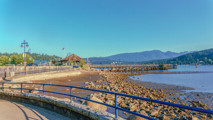 Public pier at Burrard Inlet viewed from nearby beach stroll on a hot summer day with mountains in background