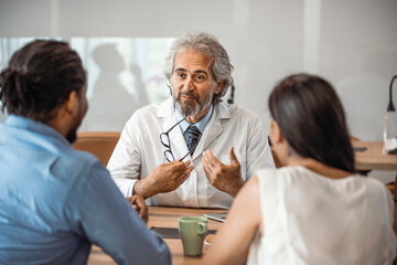 Obraz premium Male doctor offering medical advices to a young couple in office. Husband and wife in a clinic, Beautiful young couple at pregnancy consultation period. Couple meeting medical specialist at hospital