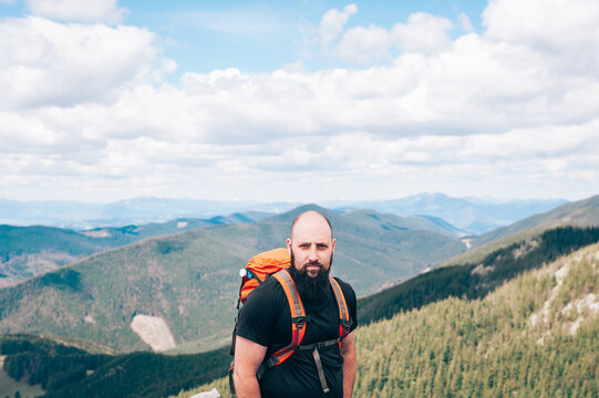 Traveler man standing on the top of mountain and looking at the camera. A handsome bald man with a beard in a black T-shirt about andorange hiking backpack stands against the backdrop of a mountain.