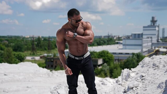 Male bodybuilder flexing muscles. African american sportsman standing shirtless showing muscles outdoors. Dark-skinned athlete in black glasses shows strong arms.