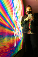 teenager dressed in black and wearing a Caribbean shirt, looking at her smart phone leaning against a bright fluorescent billboard.